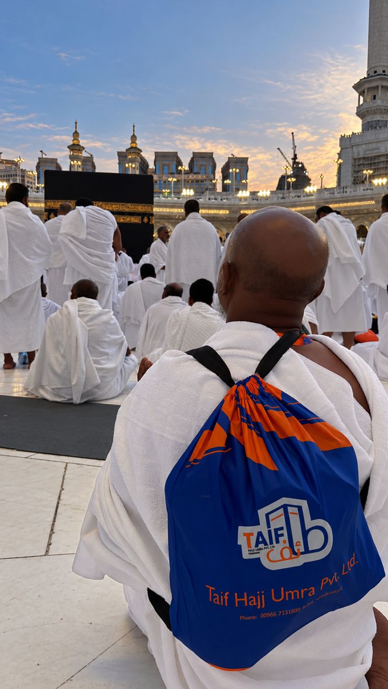 A Taif pilgrim in Ihram sitting before the Ka'bah at Masjid al-Haram, wearing the blue and orange Taif bag